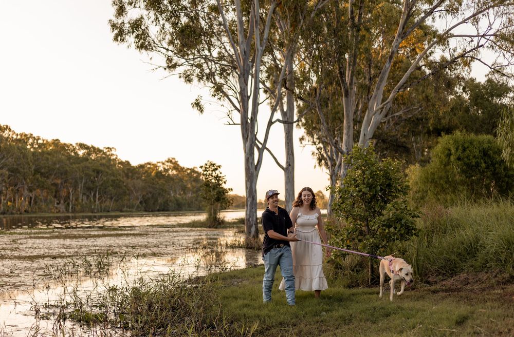 Couple walking dog along a scenic river bank at golden hour surrounded by tall eucalyptus trees.