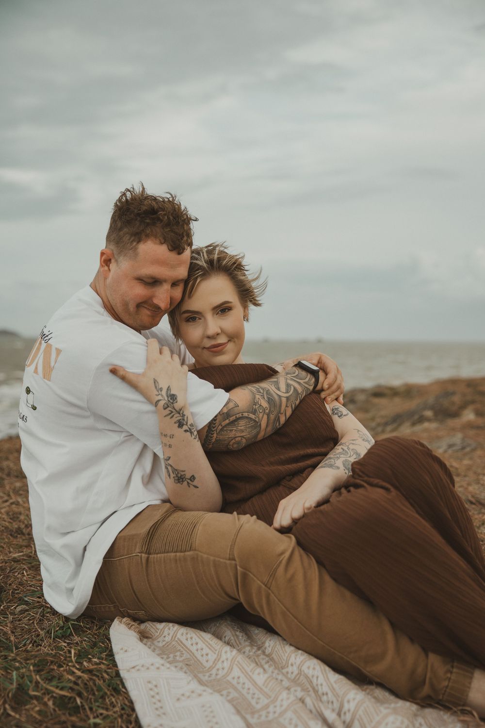 Couple with tattoos sitting on a blanket by the ocean, embracing romantically on a cloudy day.