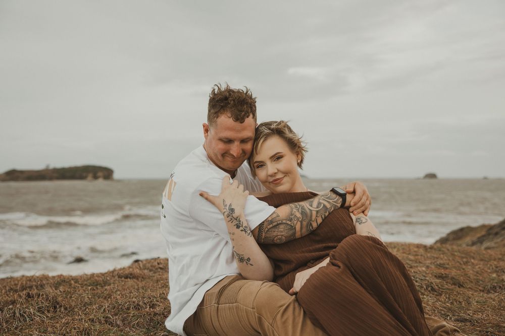 Couple embracing on rocky coastal shore with ocean waves, sitting together in casual earth-toned outfits on overcast day.