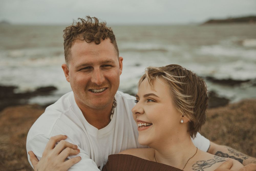 Happy couple smiling together on a rocky beach with ocean waves in the background.