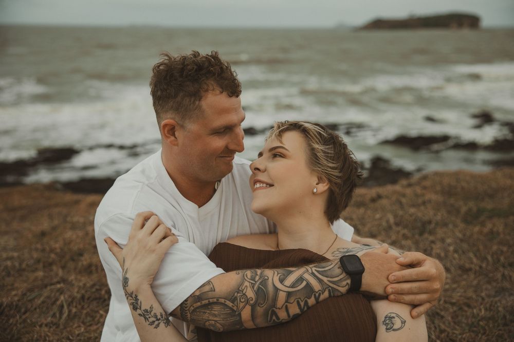 Couple embracing and smiling at each other on a rocky ocean shore, man in white tee, woman with tattoos.