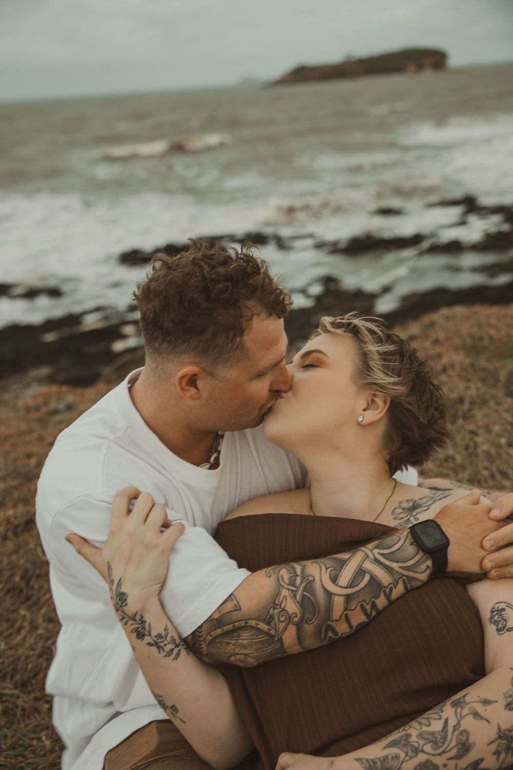 Tattooed couple sharing a romantic kiss while sitting on a rocky coastal cliff with ocean waves in the background.