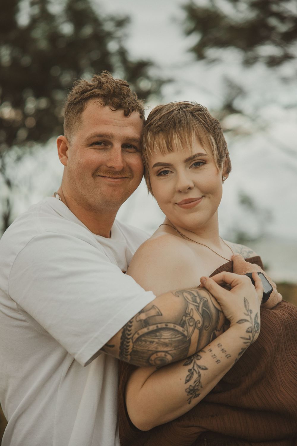 A couple embraces outdoors, woman with short hair and tattoos wearing brown top, man in white tee smiling.