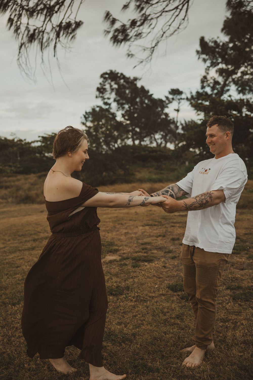 Couple holding hands and dancing barefoot in a grassy field with trees, wearing casual brown dress and white tee.