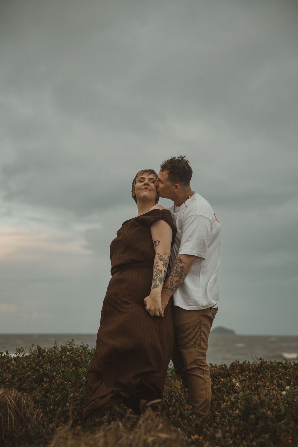 Couple sharing a tender kiss on a coastal cliff, woman in brown dress with tattoos, moody cloudy sky backdrop.