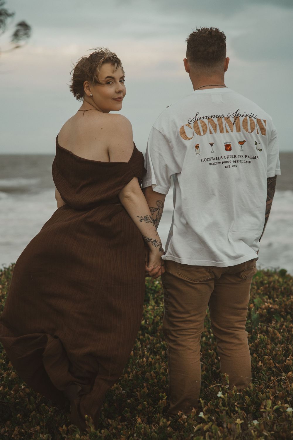 Couple holding hands by the ocean, woman in brown off-shoulder dress, man in white Common t-shirt.