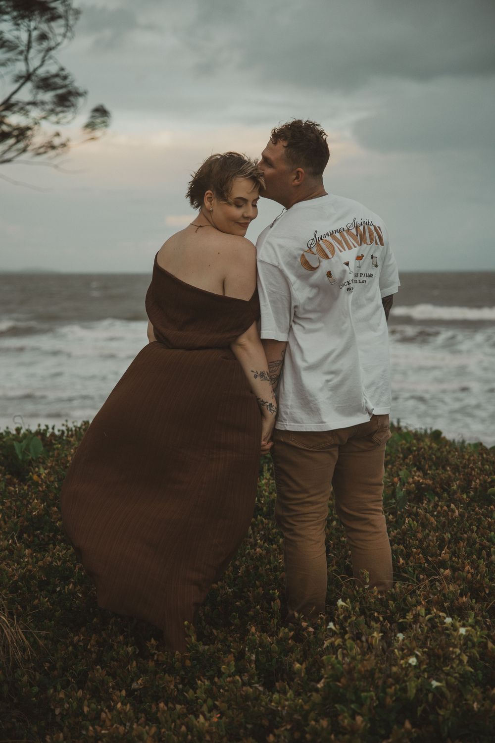 Couple sharing a romantic moment by the ocean, man kissing woman's cheek, wearing brown dress and white graphic tee.