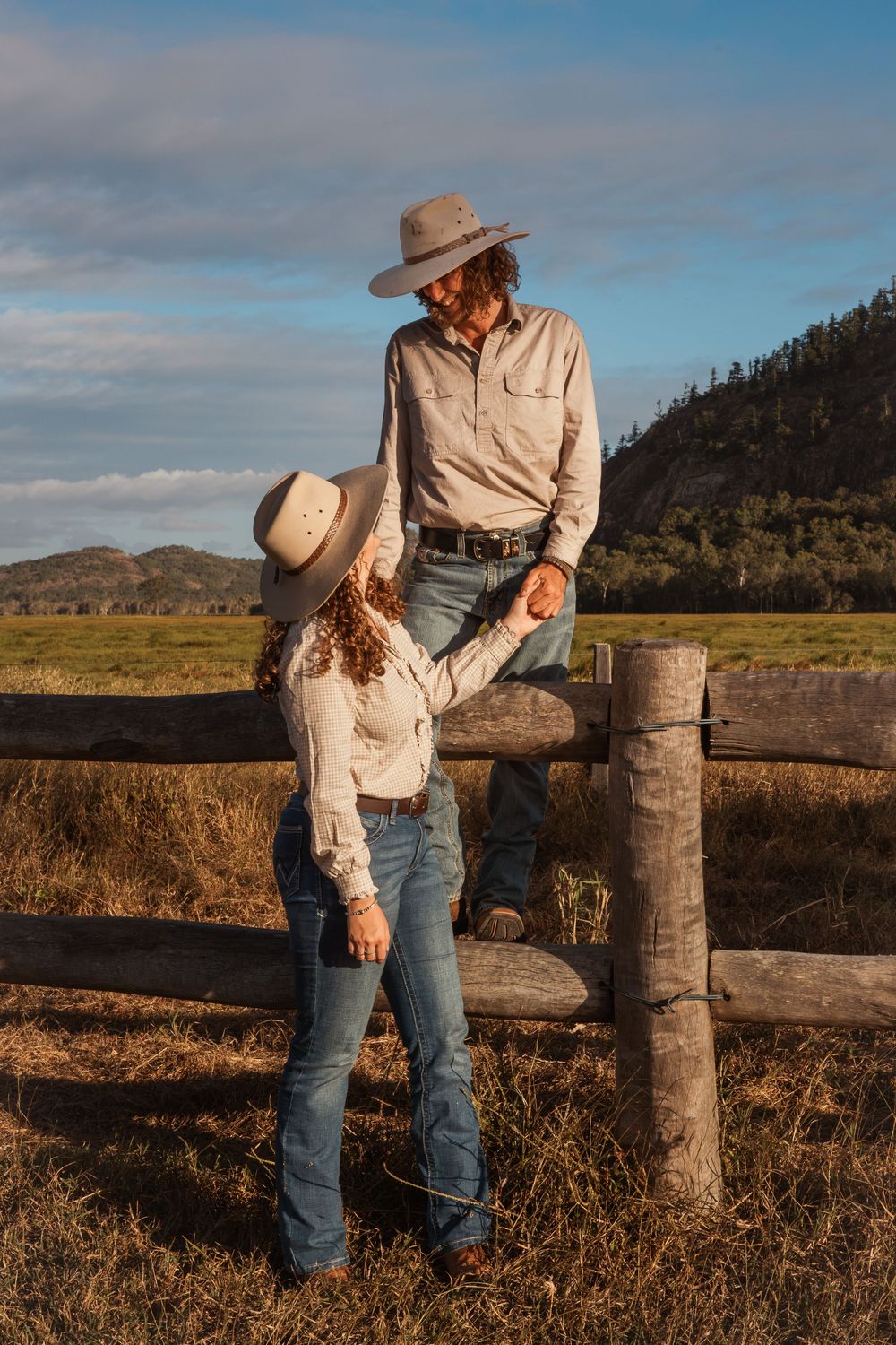 A couple in cowboy hats and jeans hold hands by a wooden fence in a sunny rural landscape.