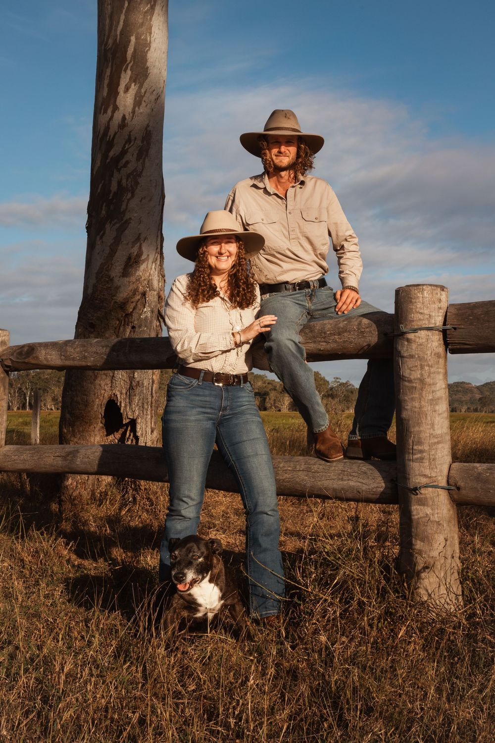 Two farmers in wide-brim hats lean on a rustic wooden fence with their dog in a sunlit rural Australian landscape.