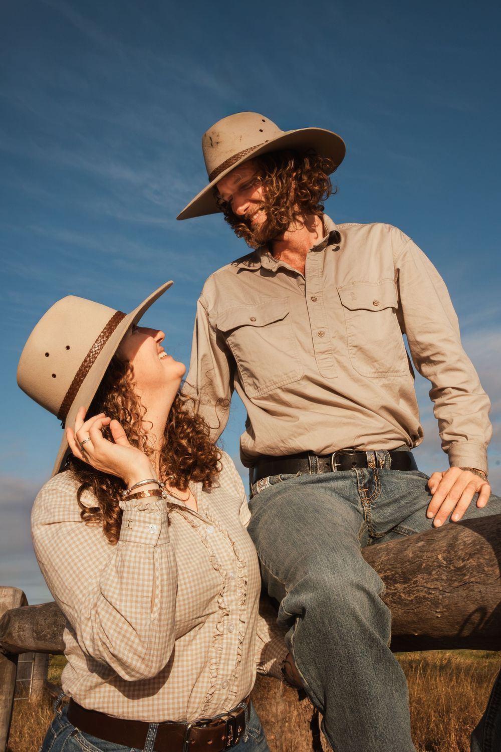 A cowboy and cowgirl couple in wide-brim hats share a tender moment by a rustic fence under a blue sky.