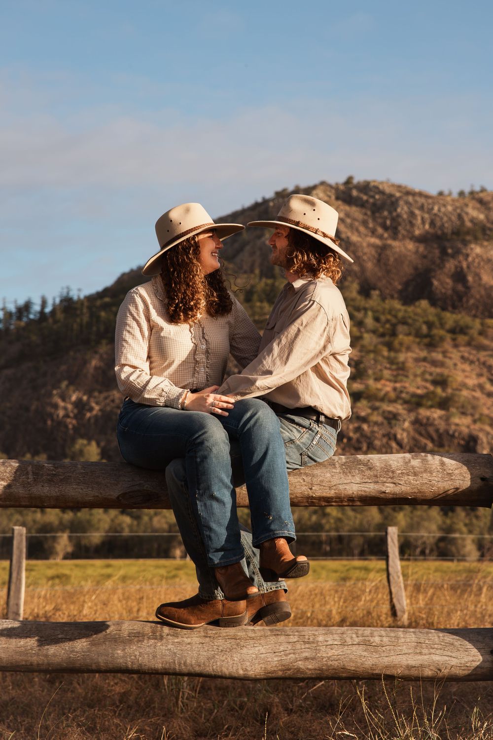 Two people in cowboy hats and jeans sit on a wooden fence, smiling at each other with mountains in the background.