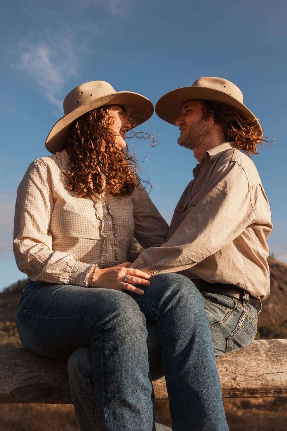 Couple in cowboy hats and jeans sitting on a fence, smiling at each other under a blue sky.