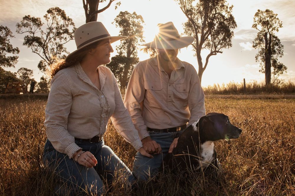 Two women in cowboy hats kneel in a golden field at sunset with a dog, surrounded by trees.