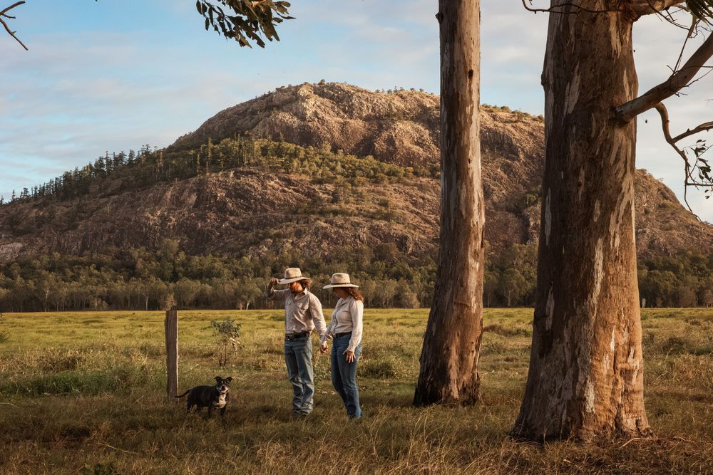 Two farmers in cowboy hats chat by a fence with their dog, set against a rocky mountain and tall gum trees in a rural field.