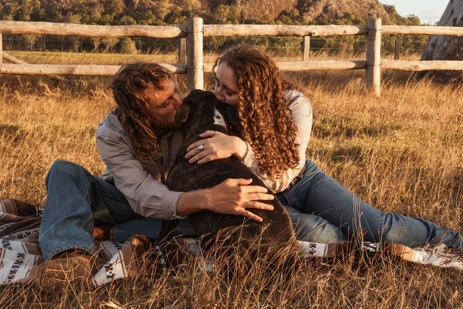 Couple with curly hair sitting in golden grass field, hugging a black dog near a wooden fence on a sunny day.