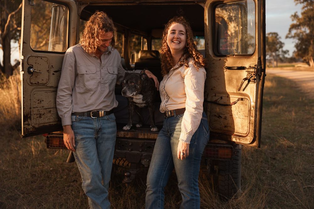 Two people and a dog pose beside a rustic vintage truck in a golden sunlit rural field.