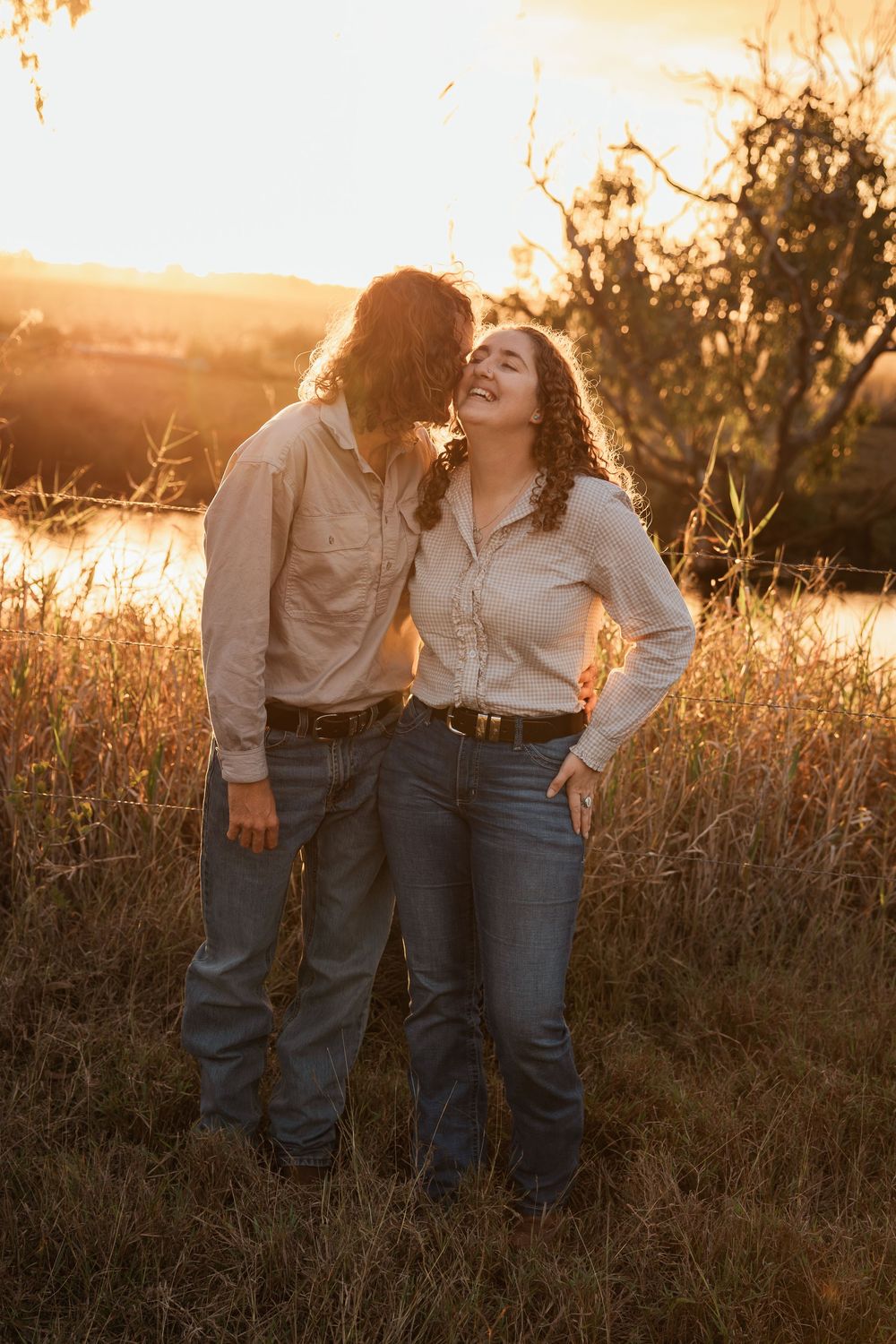 Couple in matching beige shirts and jeans embracing at golden sunset in a field near a river.
