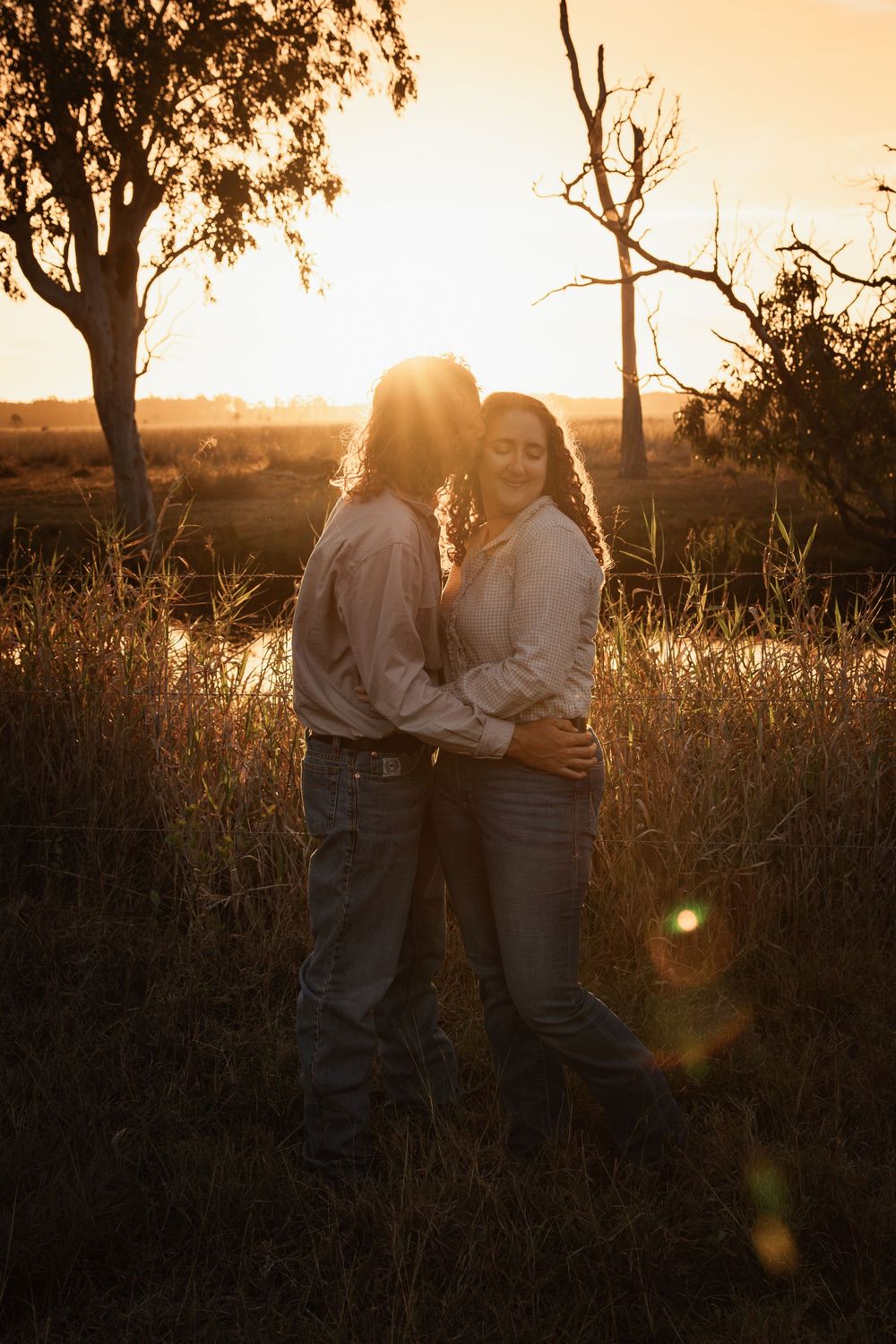 Two women embrace in a golden sunset field, surrounded by tall grass and trees near a tranquil pond.