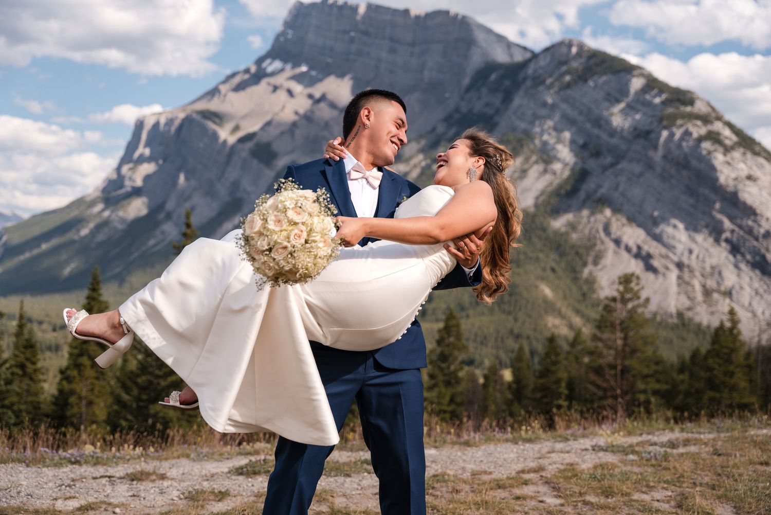 Groom lifting bride in white dress with mountain backdrop at Tunnel Mountain, Banff wedding, bouquet in hand.