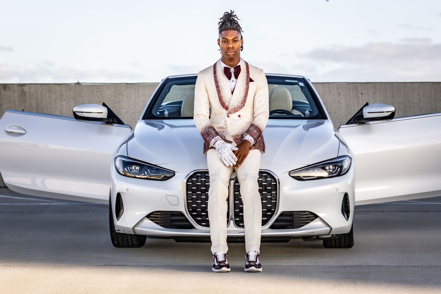 A stylish man in a white suit poses confidently in front of a sleek silver BMW convertible outdoors.