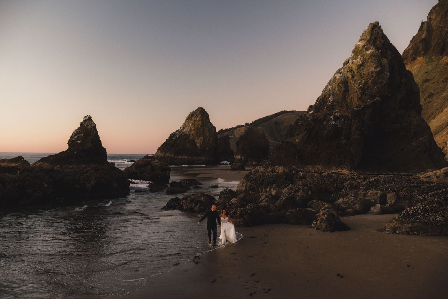 Bride and groom hold hands near a dramatic rocky sea cave on a pebble beach during their elopement.