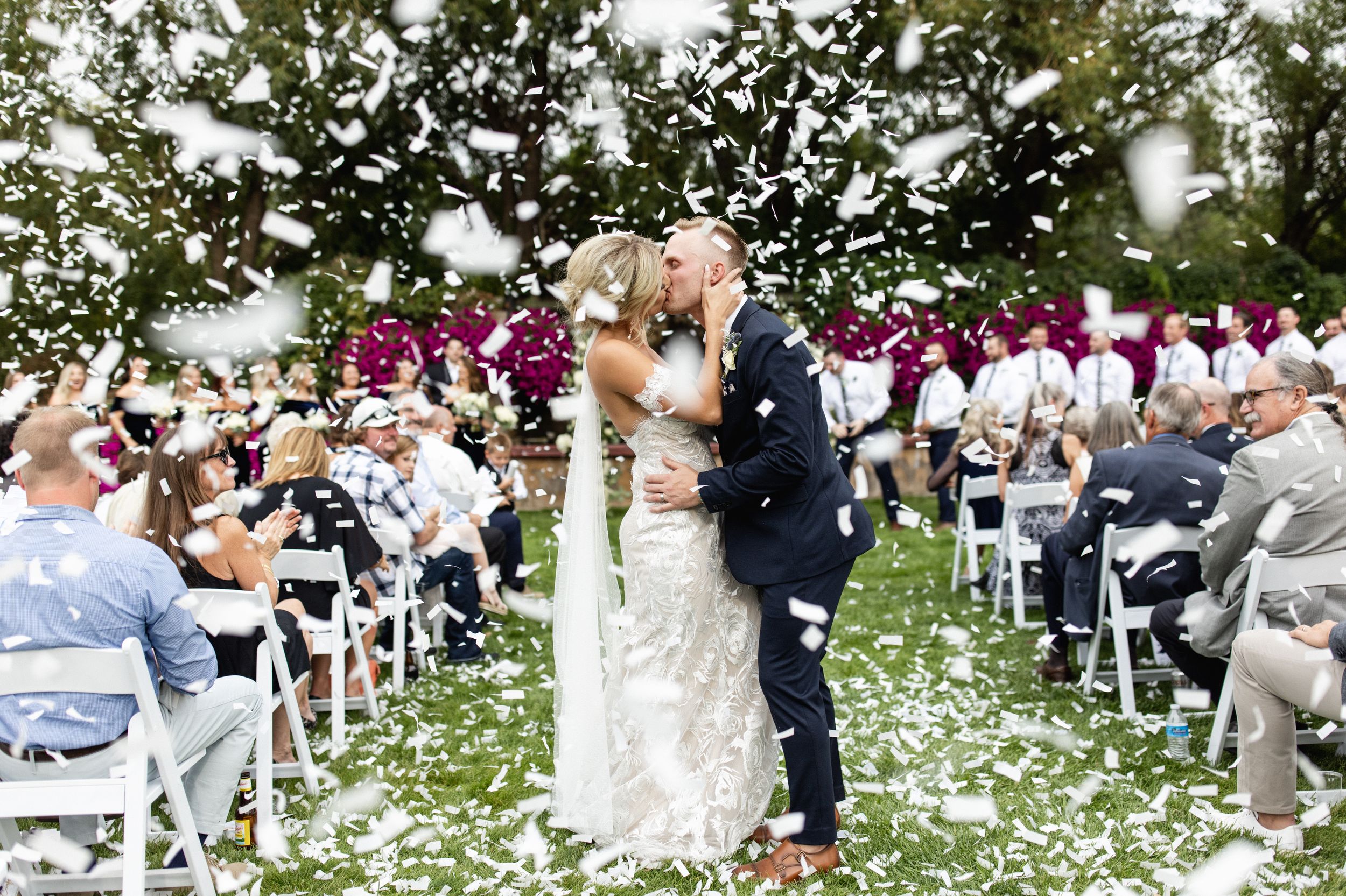 Bride and groom share a kiss during outdoor wedding ceremony with confetti falling and guests seated in white chairs.