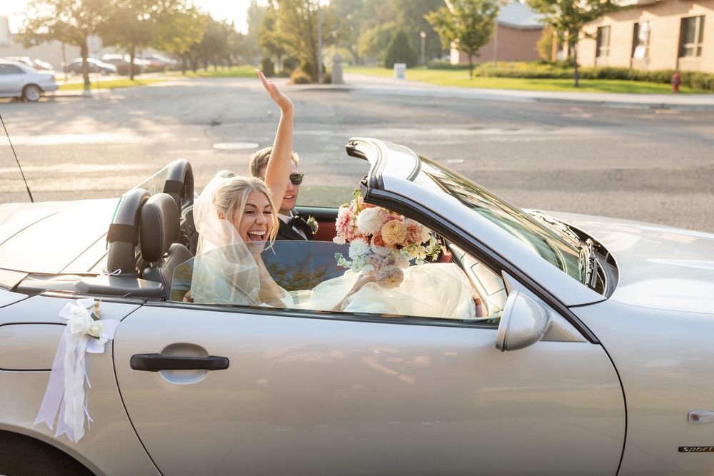 Joyful bride in white dress celebrating in silver convertible car with raised arm on sunny suburban street.
