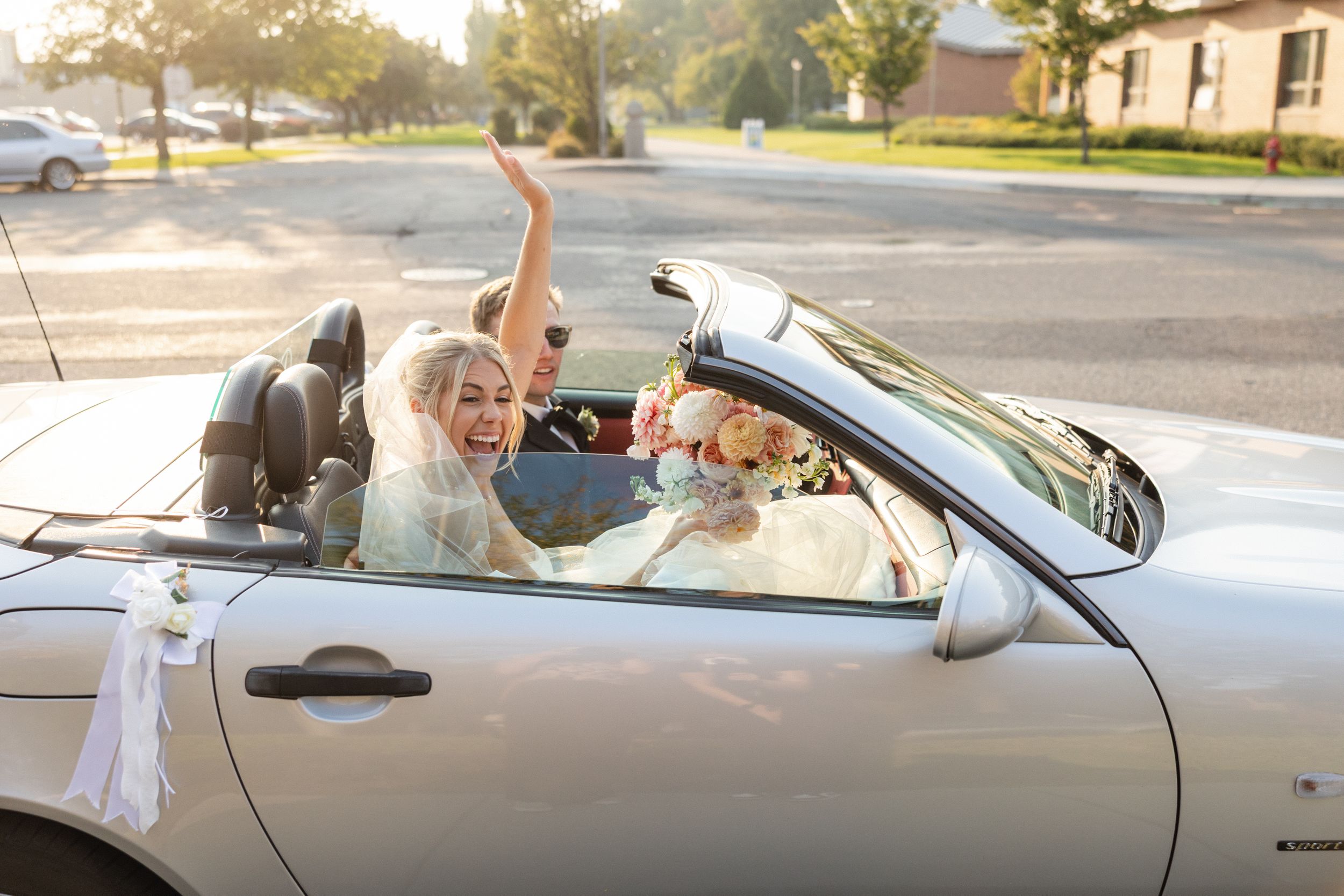 Joyful bride in white dress celebrating in silver convertible car with raised arm on sunny suburban street.