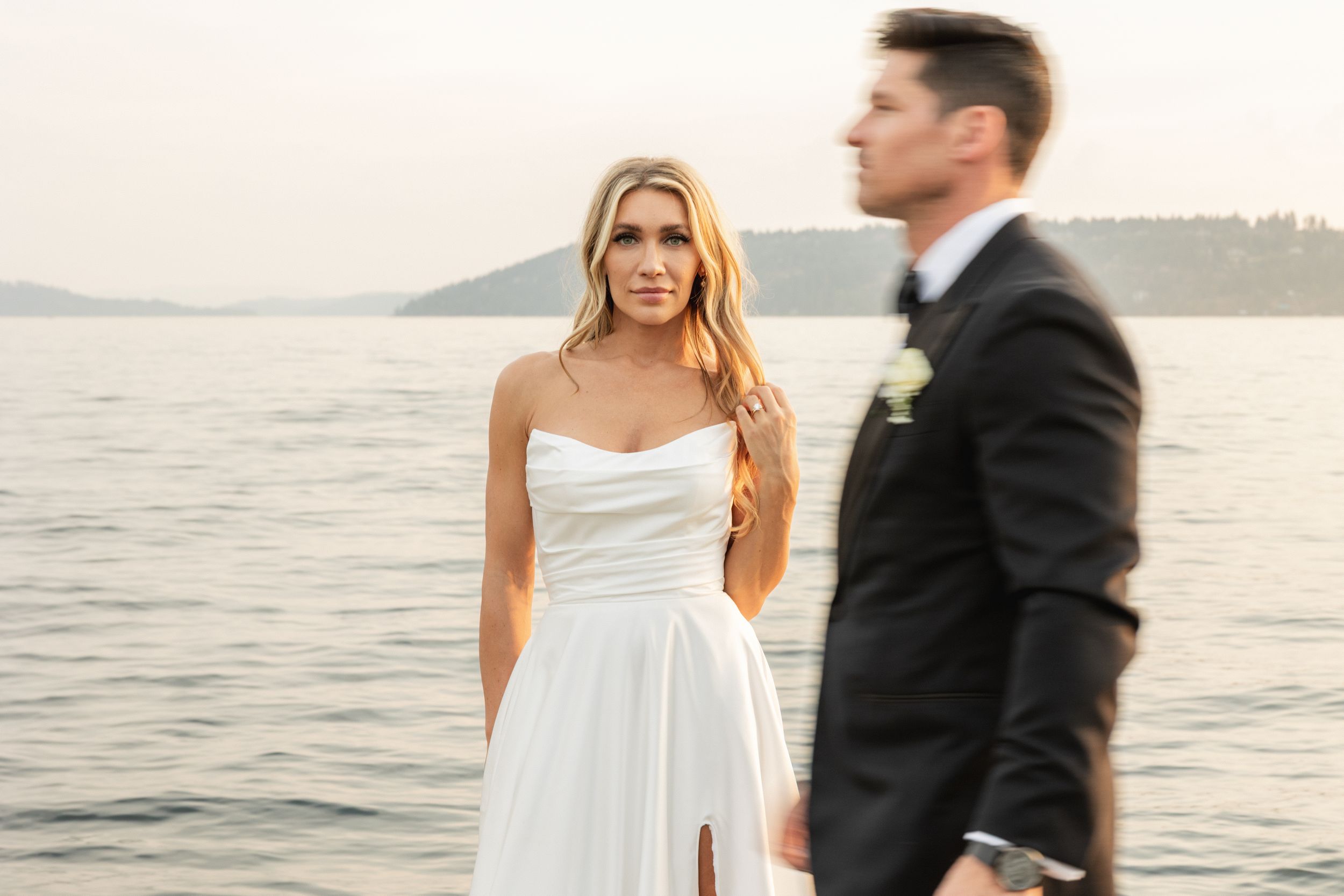 Bride in white strapless gown stands by waterfront as groom in black suit walks past during outdoor wedding.