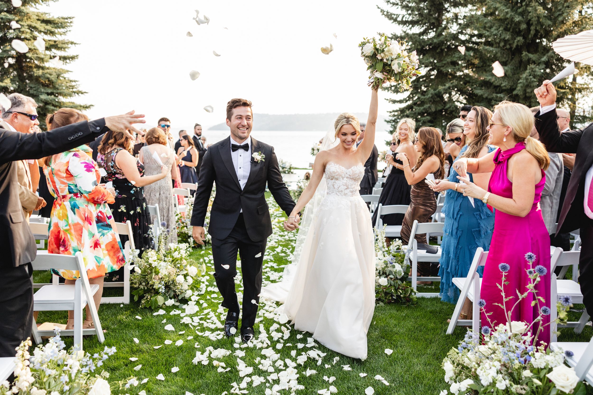 Newlyweds walk down flower-petal aisle as guests celebrate with confetti at elegant outdoor lakeside wedding ceremony.
