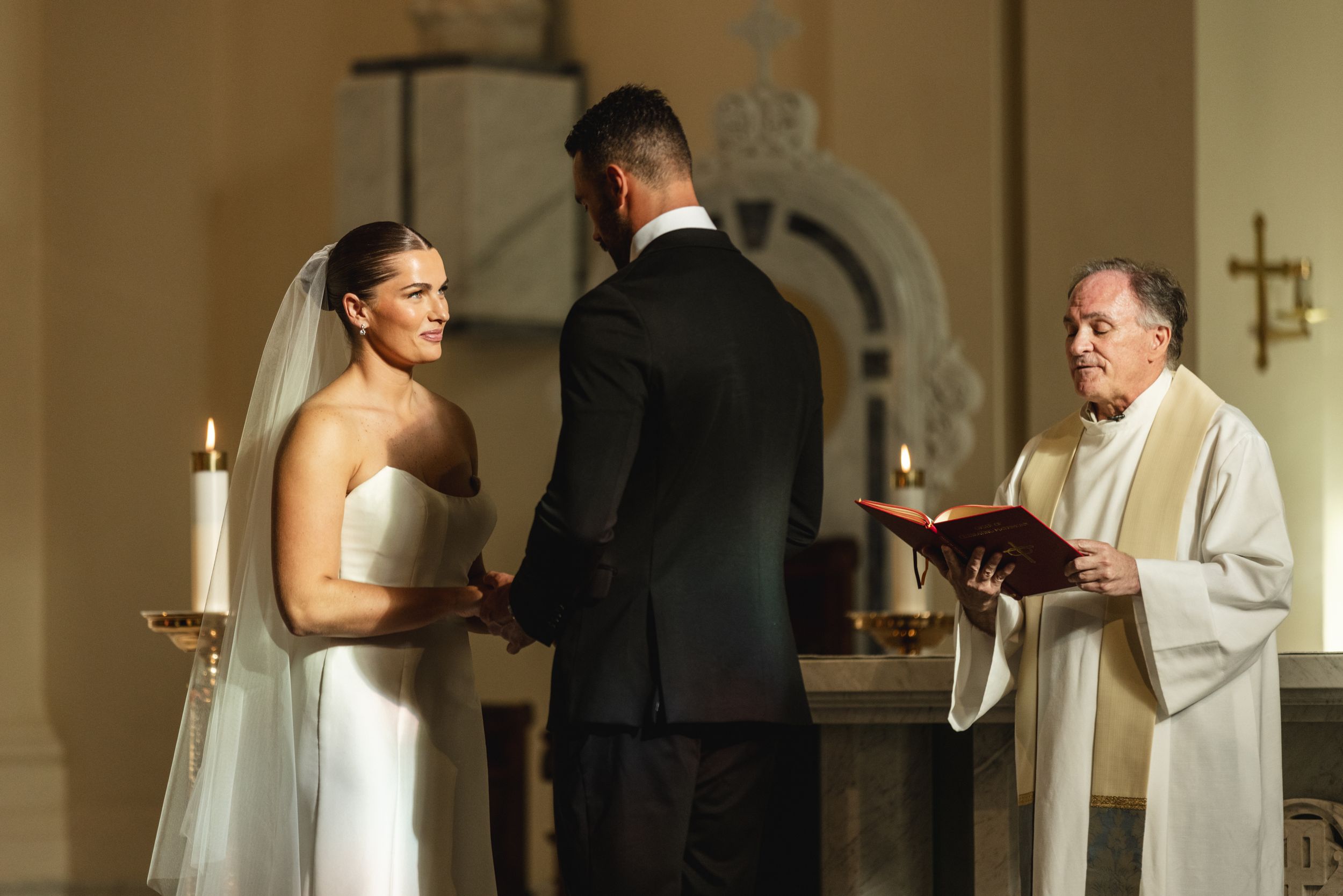 Bride and groom exchange vows at church altar with priest reading from red book during wedding ceremony.
