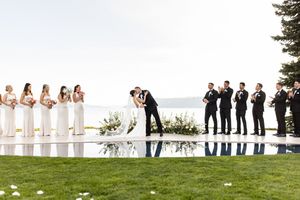 Bride and groom kiss at outdoor wedding ceremony beside reflective pool with bridesmaids and groomsmen, lake view backdrop.