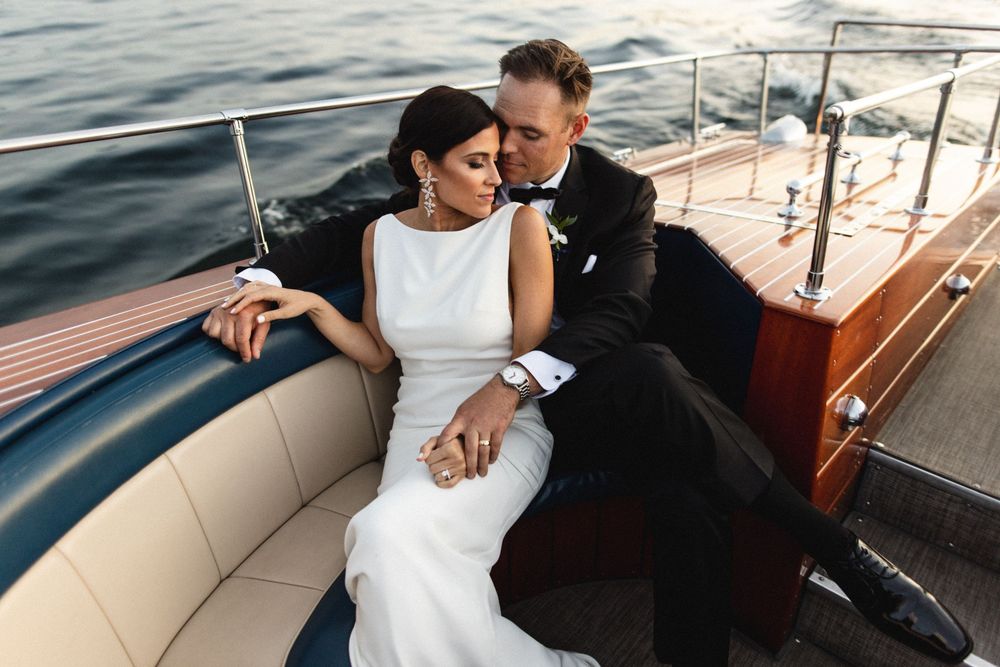 Elegant bride and groom in wedding attire sitting closely together on a luxury wooden boat on calm water.