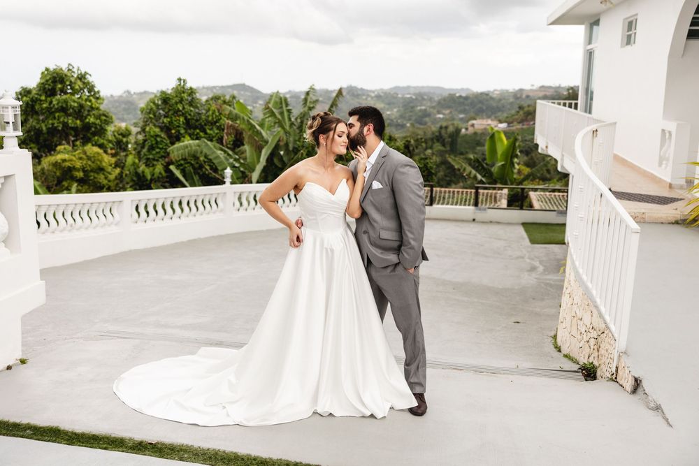 Bride and groom share a romantic moment on a white terrace with lush tropical greenery in the background.