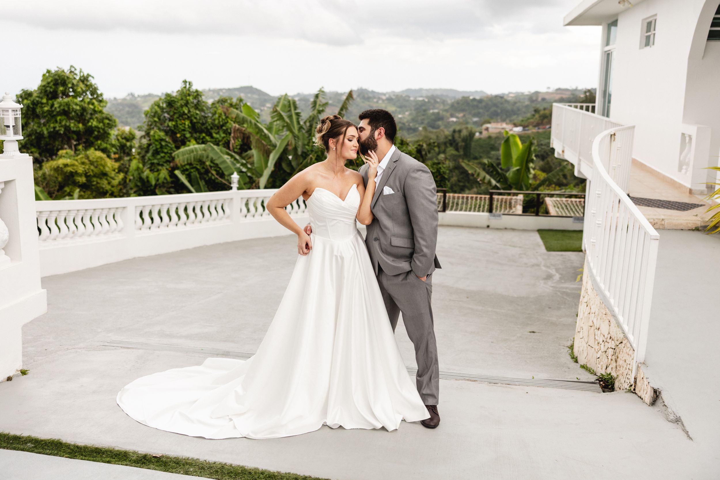 Bride and groom share a romantic moment on a white terrace with lush tropical greenery in the background.