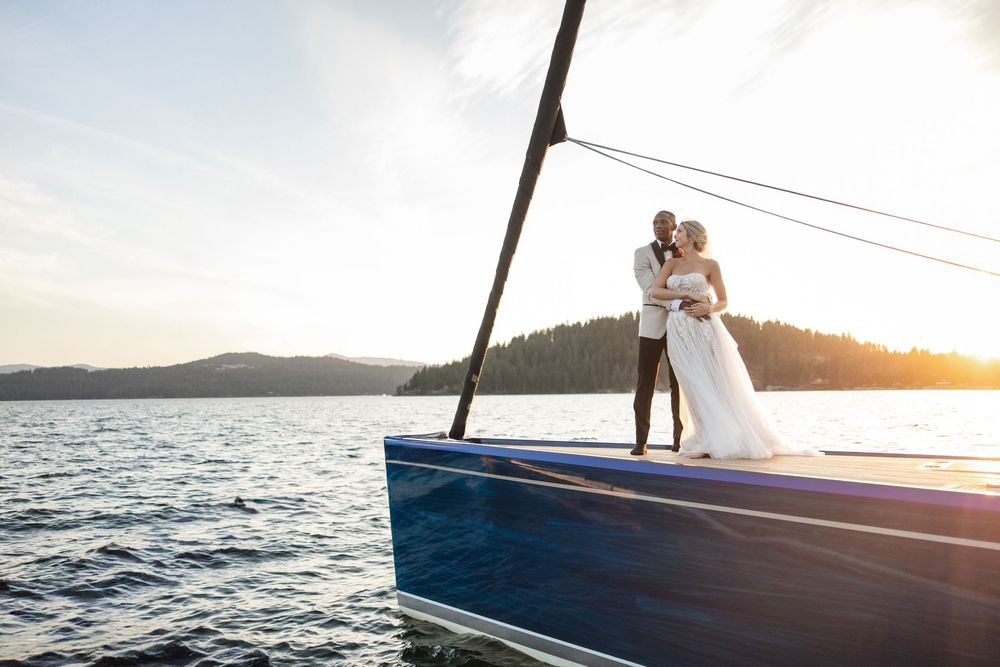 Bride and groom embracing on the bow of a sailboat at sunset with mountains and calm water in background.