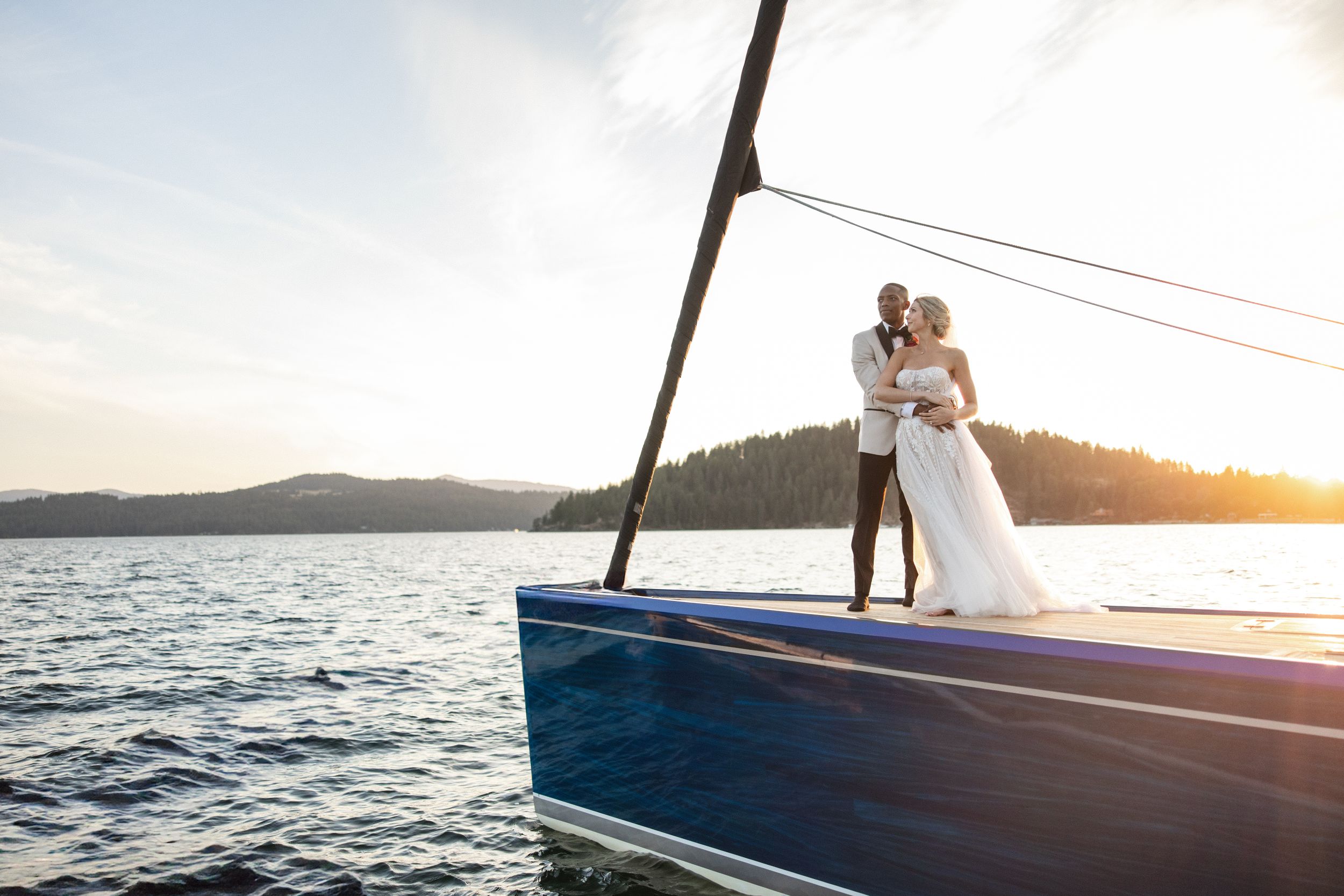 Bride and groom embracing on the bow of a sailboat at sunset with mountains and calm water in background.