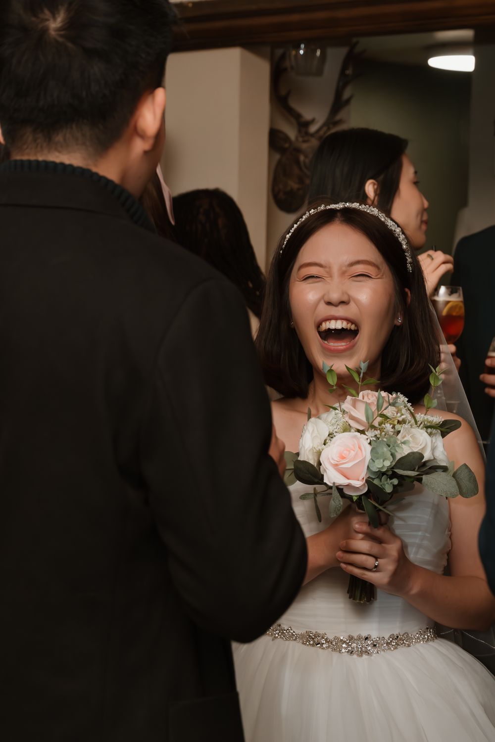 Joyful Asian bride laughing while holding a pink rose bouquet at her wedding reception in a white gown.