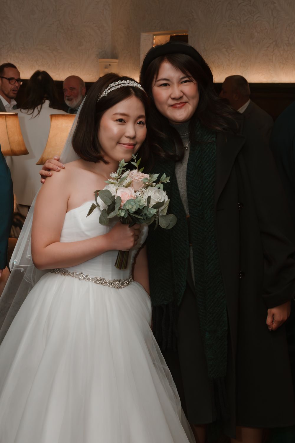 Bride in white gown holding bouquet poses with guest in green coat at wedding reception.