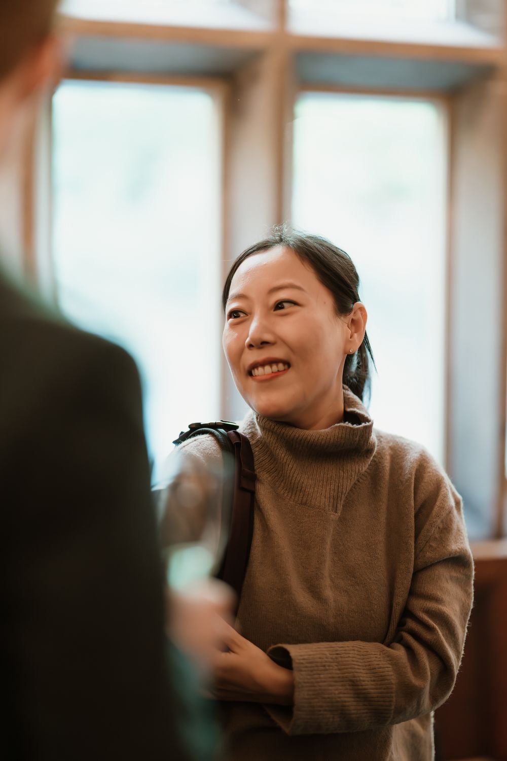 Smiling Asian woman in a brown turtleneck sweater having a conversation indoors near bright windows.