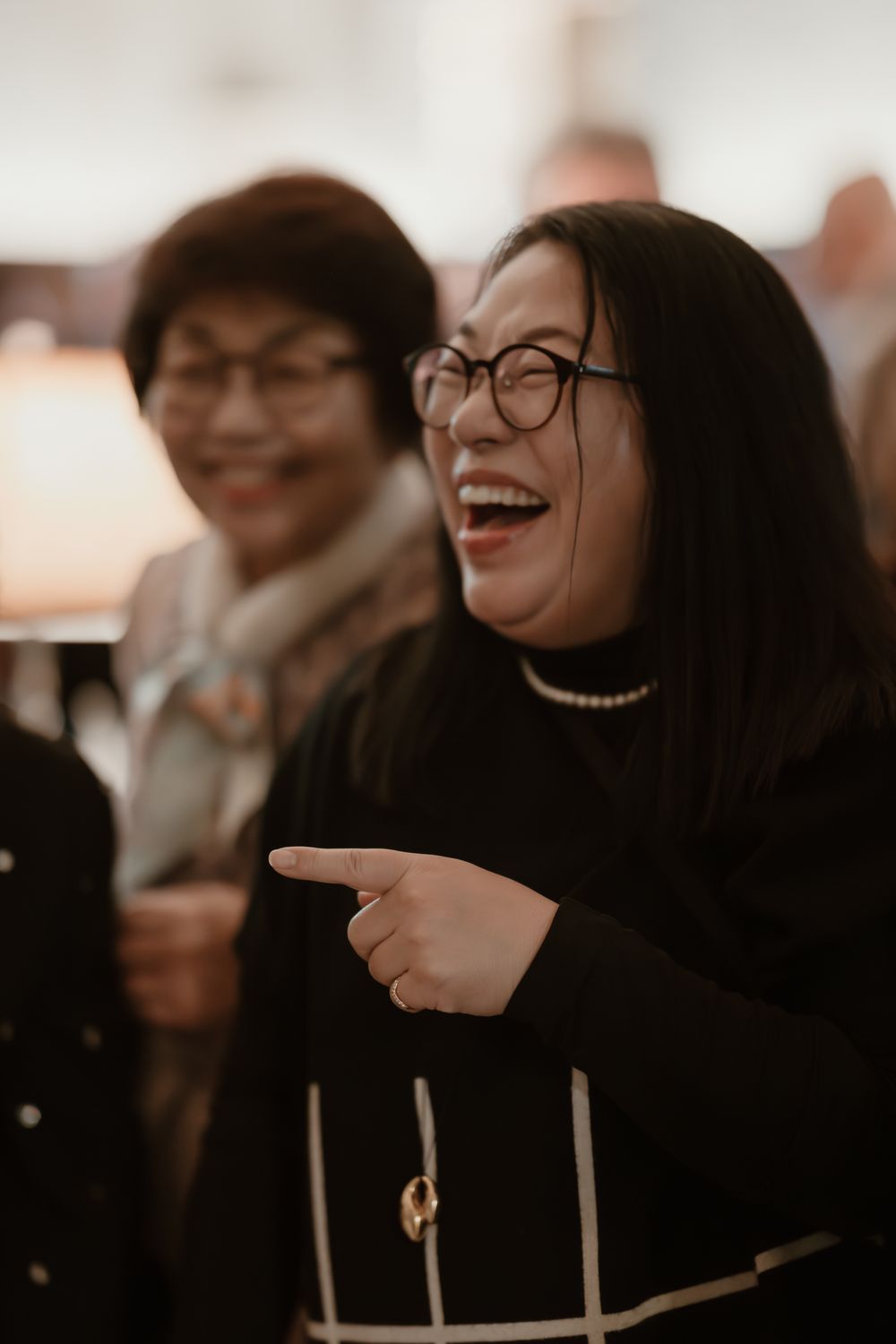 Two Asian women laughing and pointing at an event, wearing glasses and dark clothing in a warmly lit room.