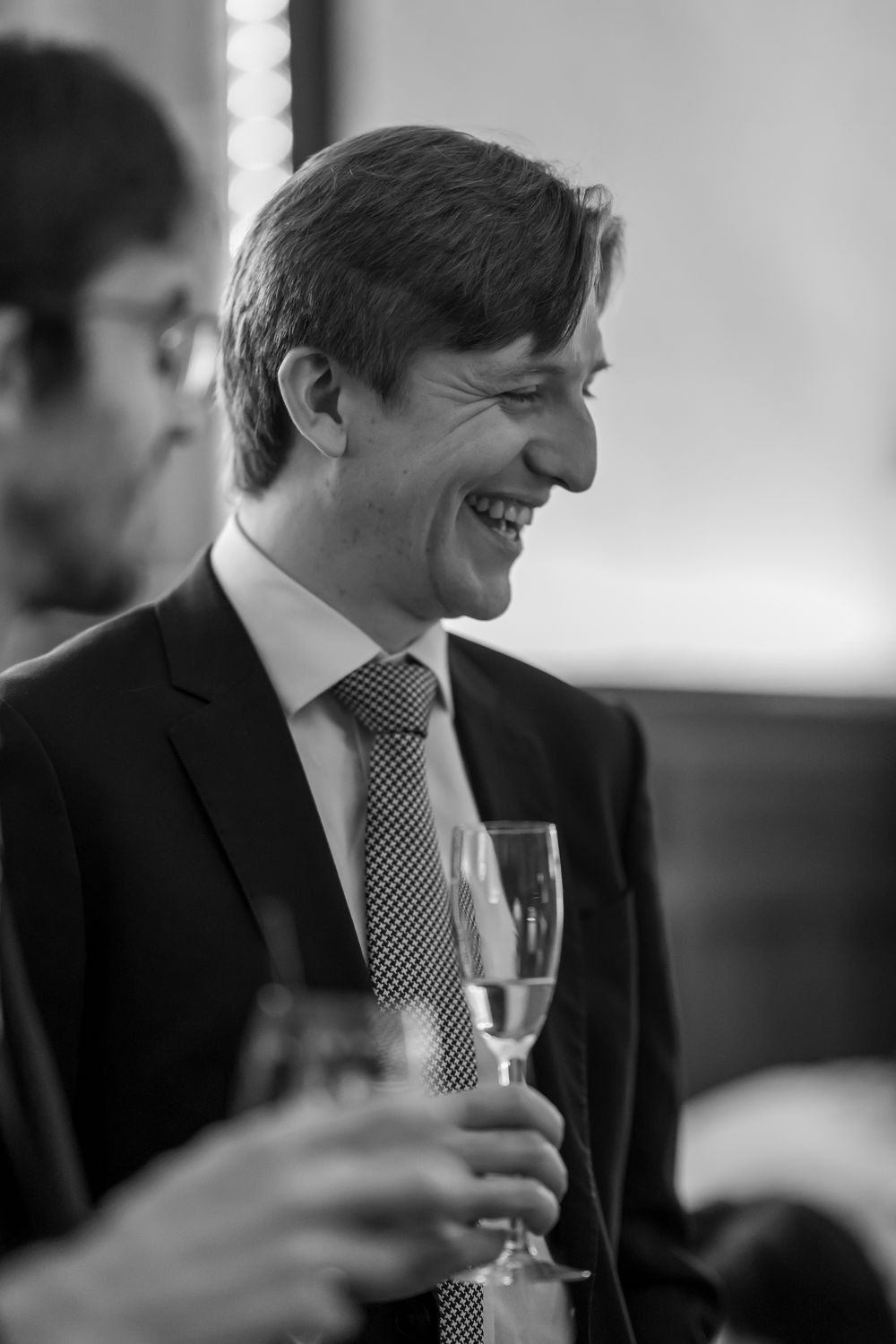 A smiling man in a suit and tie holds a champagne glass at a formal event, captured in black and white.
