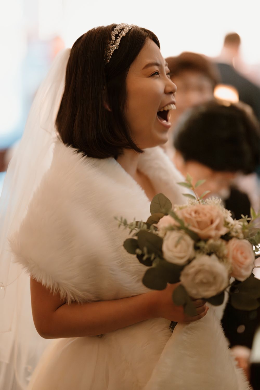 Joyful Asian bride in white gown with fur shawl and floral bouquet laughing during wedding ceremony.