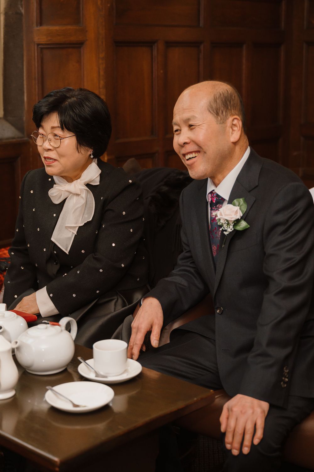 Elderly Asian couple in formal attire sitting at a table with tea cups, man smiling and wearing a boutonniere.