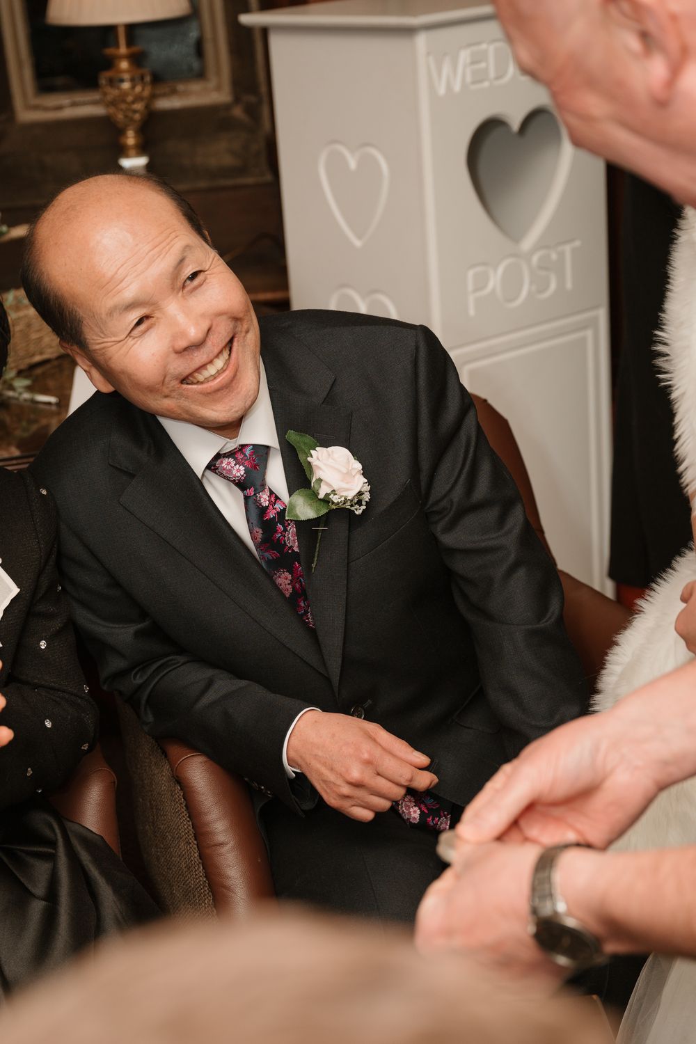 Smiling man in black suit with floral boutonniere at a wedding reception near a decorative post box.