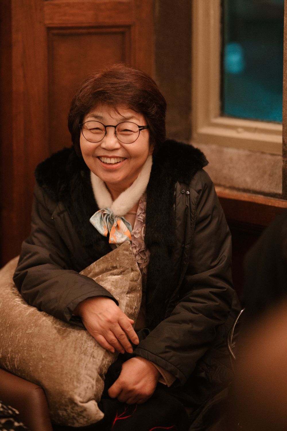 Smiling middle-aged Asian woman wearing glasses and a black winter jacket, holding a pillow indoors.