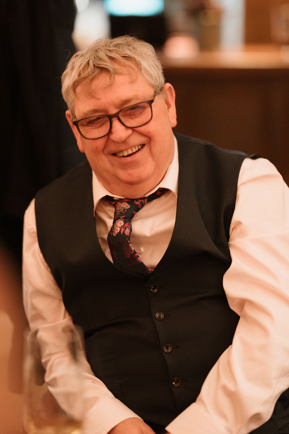 Smiling older man with glasses wearing a black vest and floral tie at a formal event.