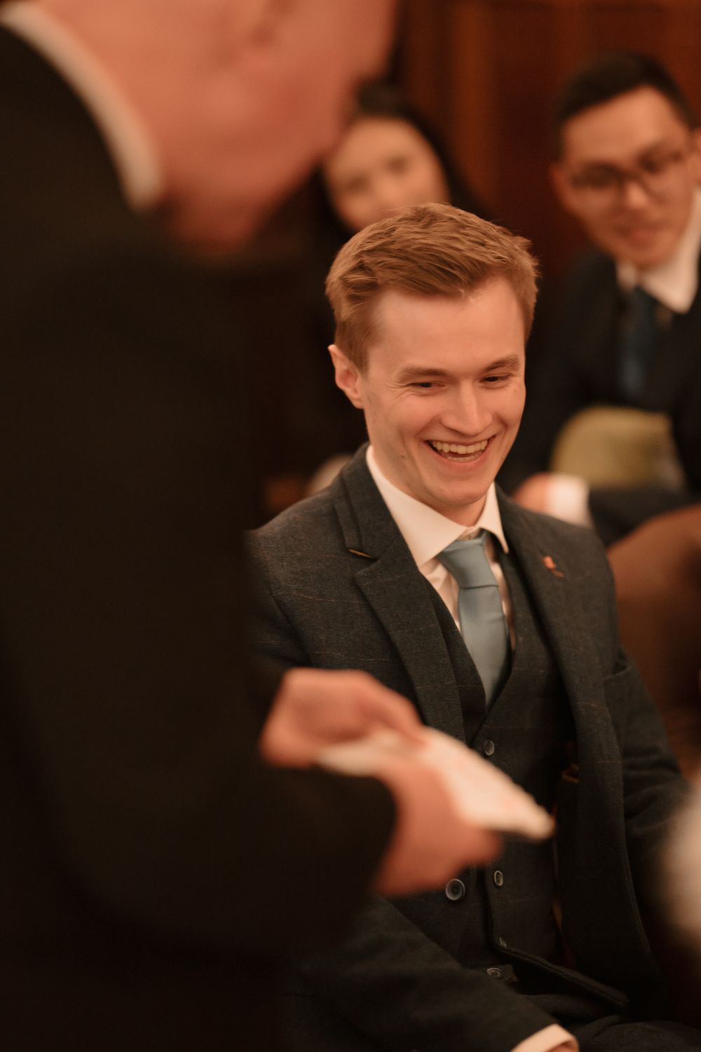 Young man in a navy suit smiling during a formal indoor event or meeting.