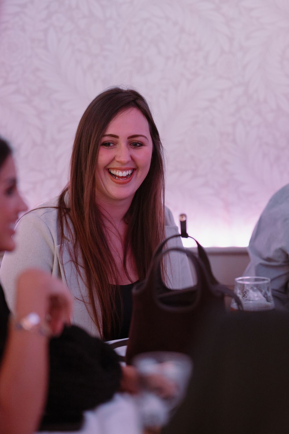 A smiling brunette woman laughs joyfully at a social gathering, seated at a table in an elegant venue.