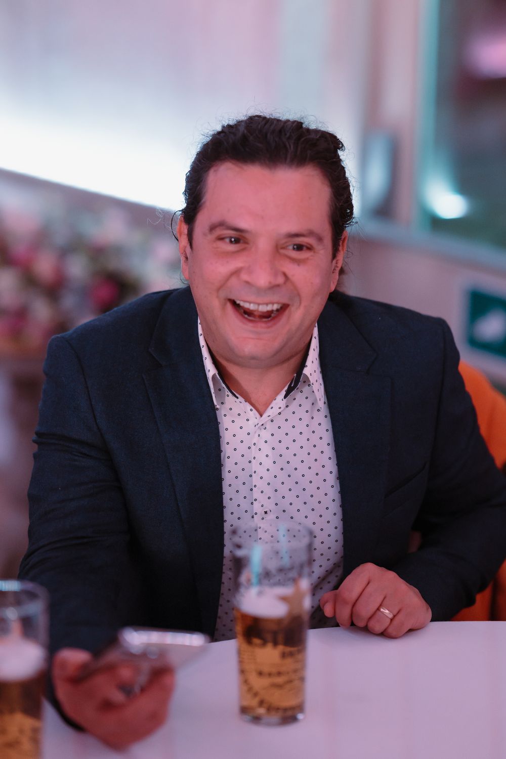 A smiling man in a dark blazer and polka dot shirt sits at a table with a glass of beer.